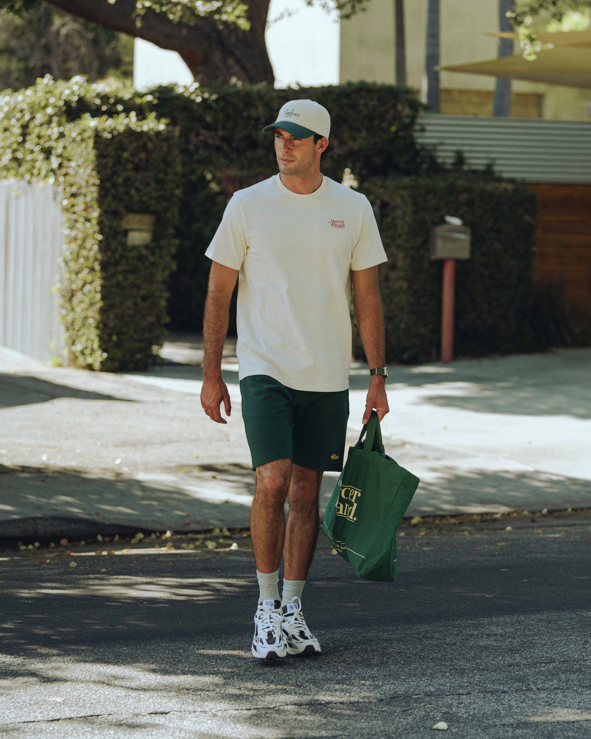 Man walking outdoors holding a green bag, wearing a white t-shirt and green shorts by the mercer brand