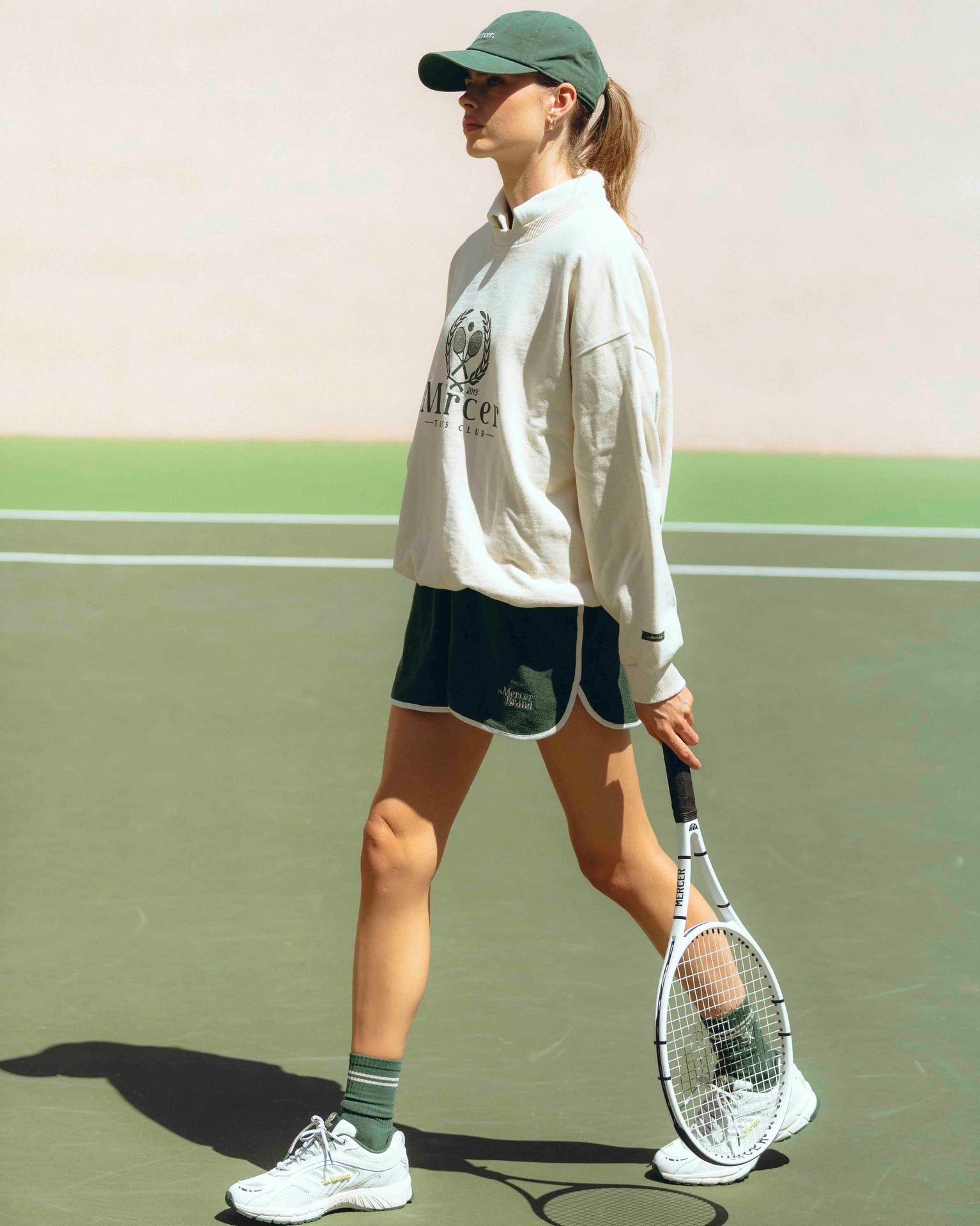 A woman is walking on a tennis court; she is wearing a Mercer cap, a Mercer sweater, and Terry shorts. Underneath, she is wearing green Mercer tennis socks paired with white Re-Run Evo sneakers.