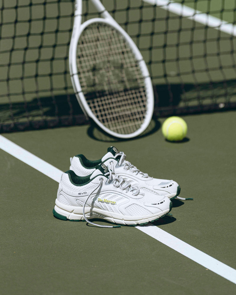 A pair of white Re-Run EVO sneakers are standing on a tennis court, viewed from the side.