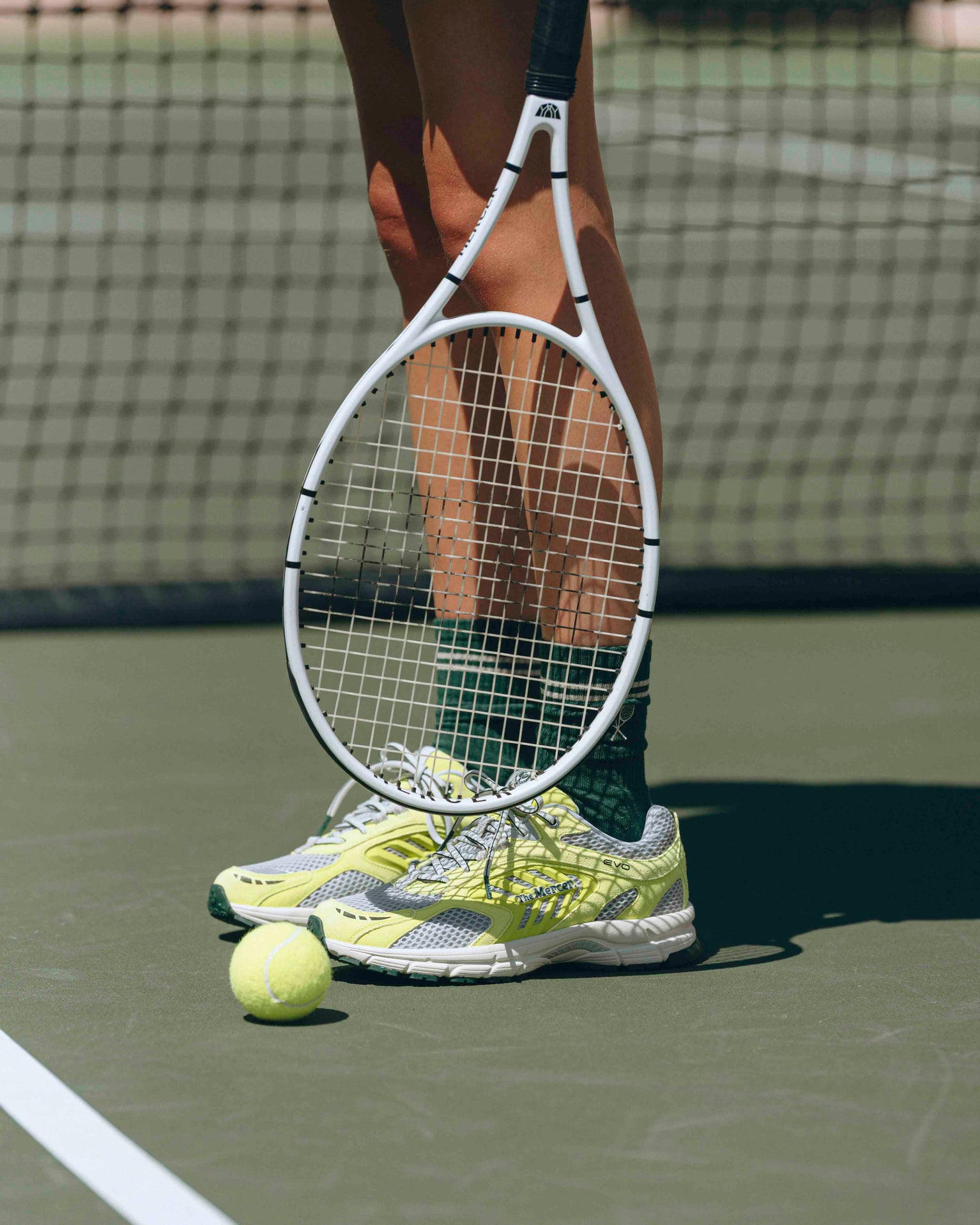 A woman is wearing The Re-Run Evo in yellow with The Mercer Tennis Socks in green. In her hand she is holding a tennisracket.