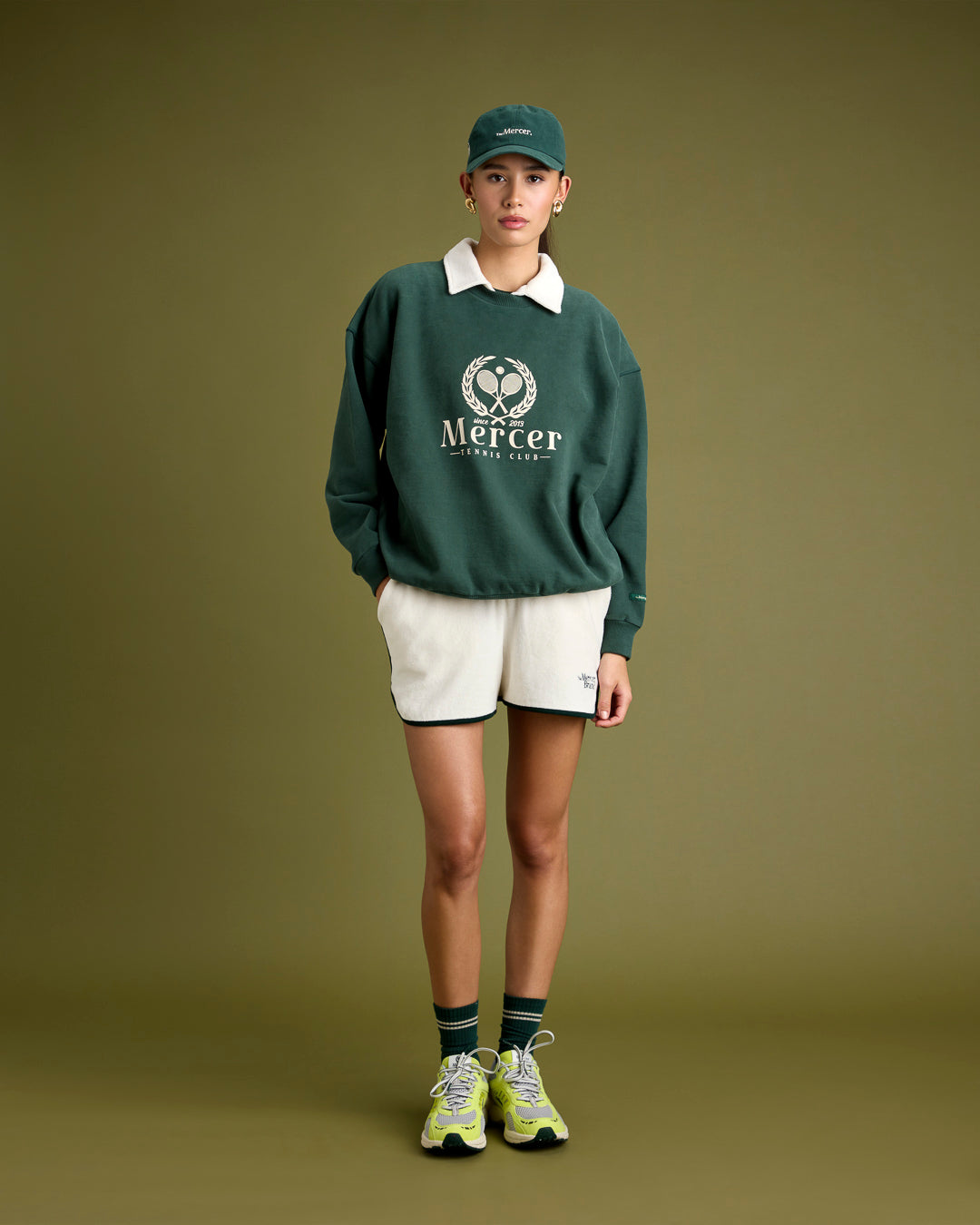 a woman is wearing The Mercer Tennis cap with the matching Tennis sweater. underneath she is wearing a The Mercer Brand tennis set with the Tennis socks and The Re-Run Evo in Yellow.