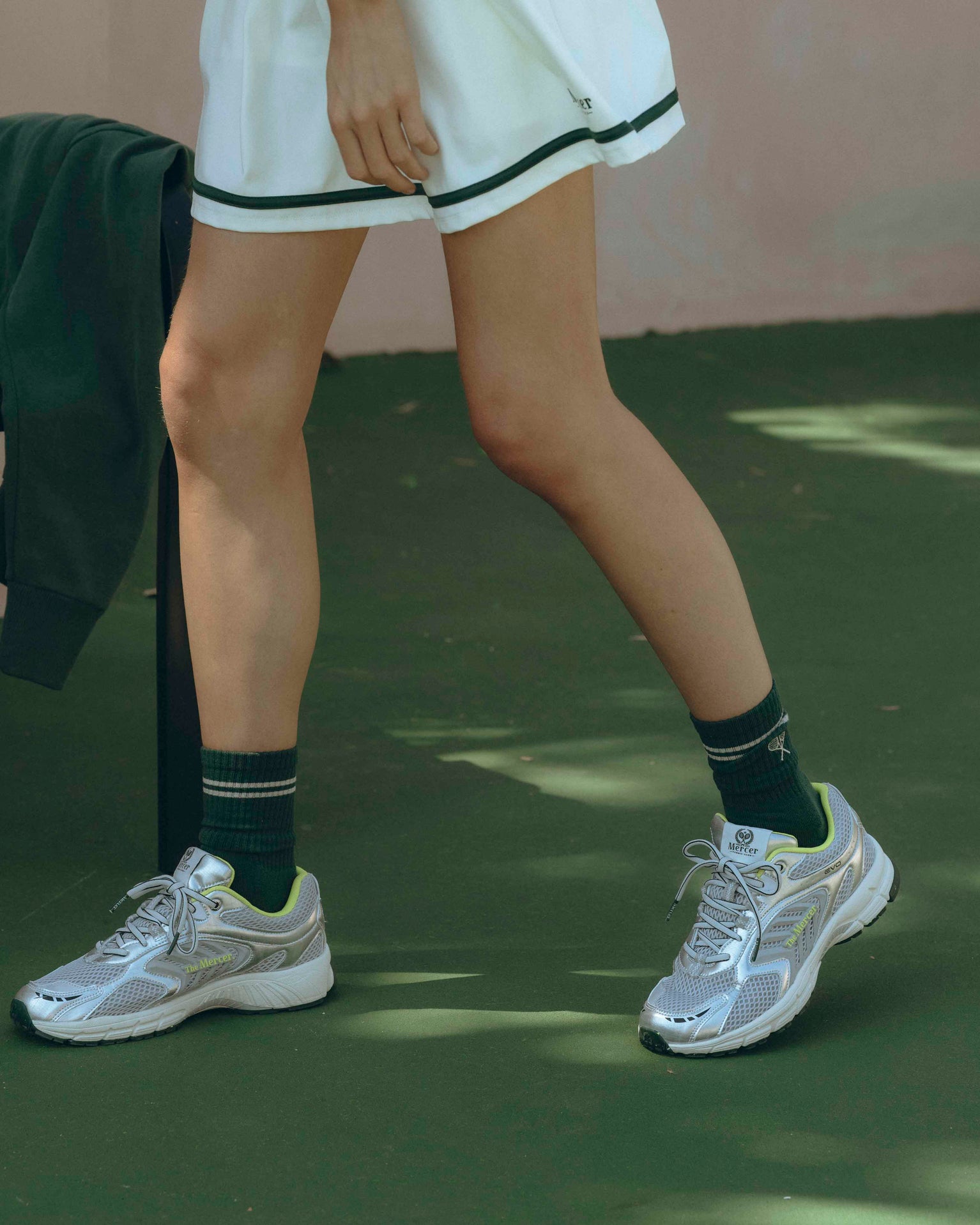 Lifestyle shot of female wearing the mercer brand re-run evo in silver on tennis court with tennis skirt and green skirt
