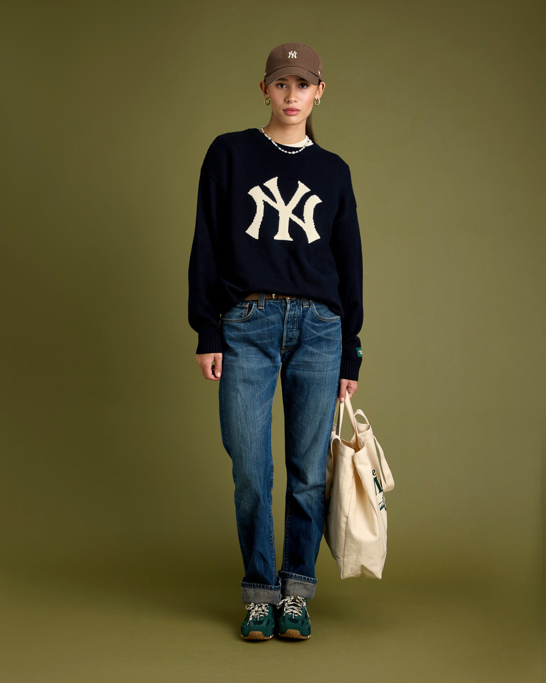 A woman wearing a navy New York Yankees knit sweater and a brown New York Yankees cap from The Mercer Brand x 47. She is also wearing The Re-Run Drive in green along with a canvas bag.