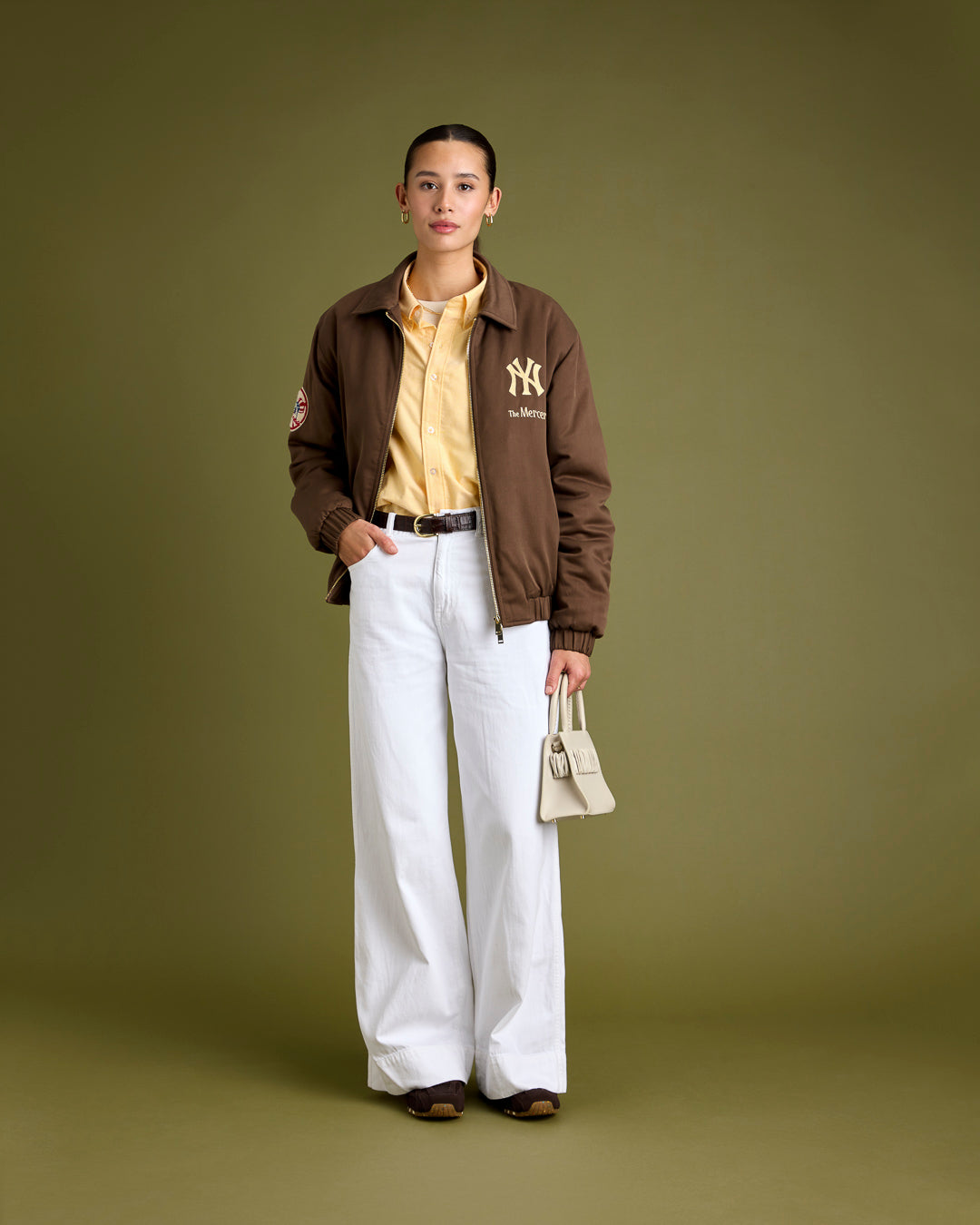 A woman is wearing a The Mercer 47 jacket in brown featuring the New York Yankees logo, paired with yellow Oxford shoes. The shoes she’s wearing with this outfit are The Re-Run Drive in brown.