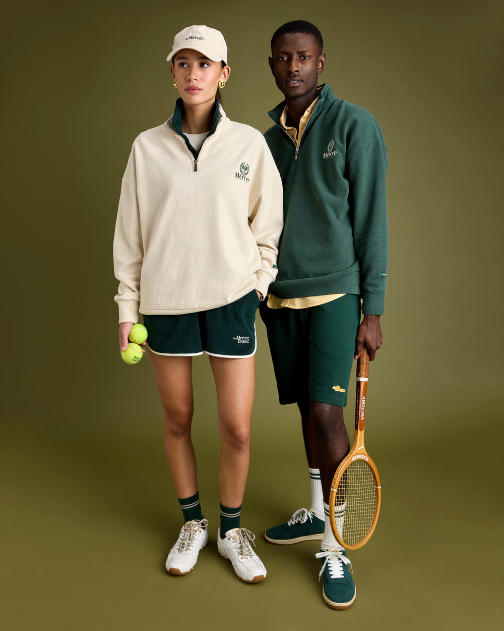 A man and a woman are both wearing a The Mercer Brans Tennis quarter zip, short and The Mercer Tennis socks. He is wearing The Re-Tennis in green and she is wearing the Re-Run Drive.