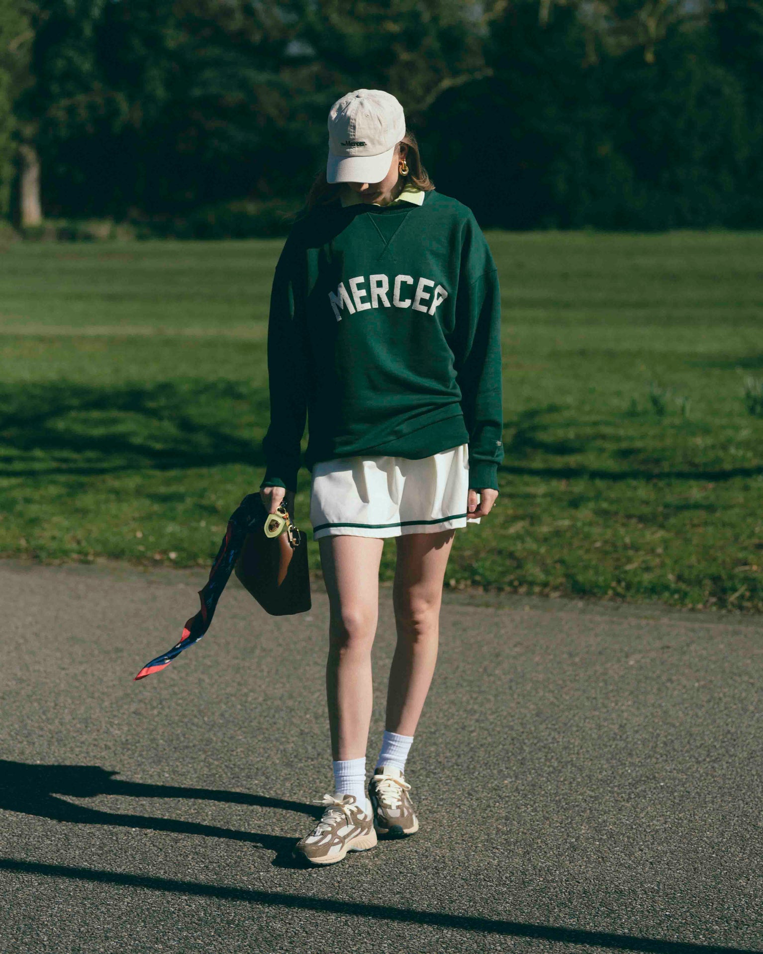 A woman is wearing The Mercer Tennis Skirt with a Mercer Sweater and cap. For shoes she is wearing The Re-Run Desert 2.0 in brown.