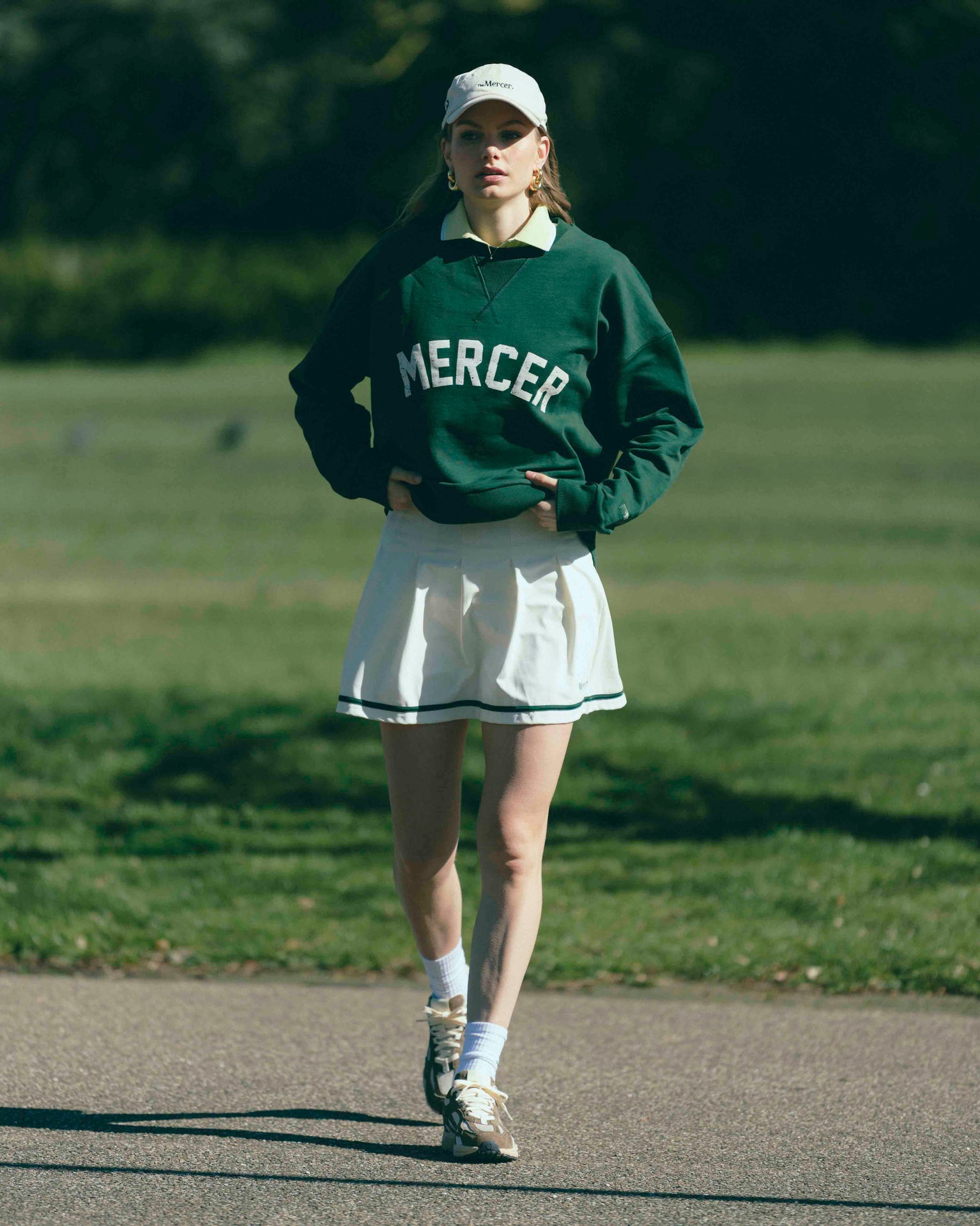 A woman is wearing The Mercer Tennis Skirt with a Mercer Sweater and cap. For shoes she is wearing The Re-Run Desert 2.0 in brown.