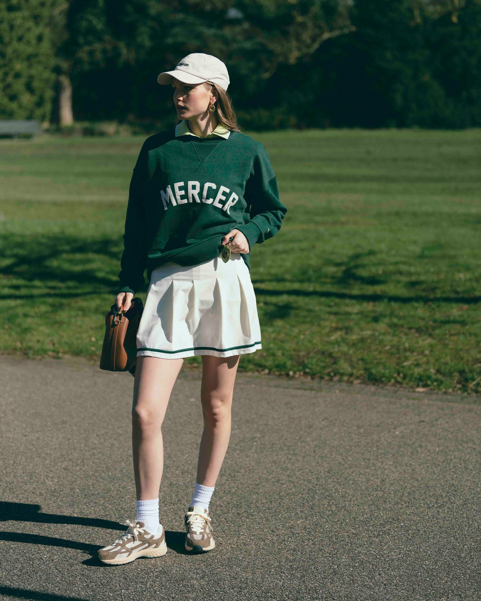 A woman is wearing The Mercer Tennis Skirt with a Mercer Sweater and cap. For shoes she is wearing The Re-Run Desert 2.0 in brown.
