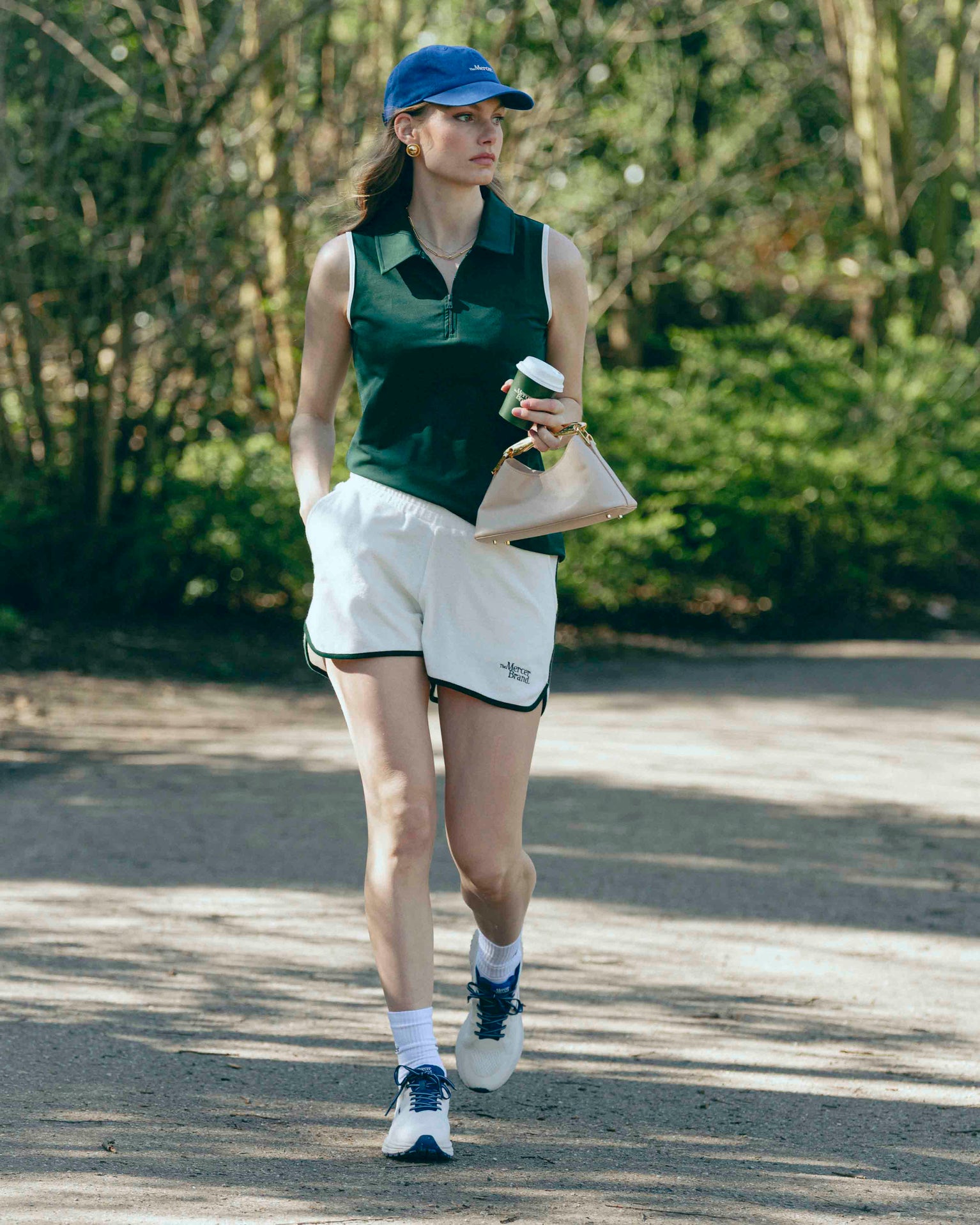 A woman is wearing the Mercer Tennis polo with The shorts and also matching the Mercer Cap with her The Mercer P22 in white/blue. In her hand she is holding her coffee to go.