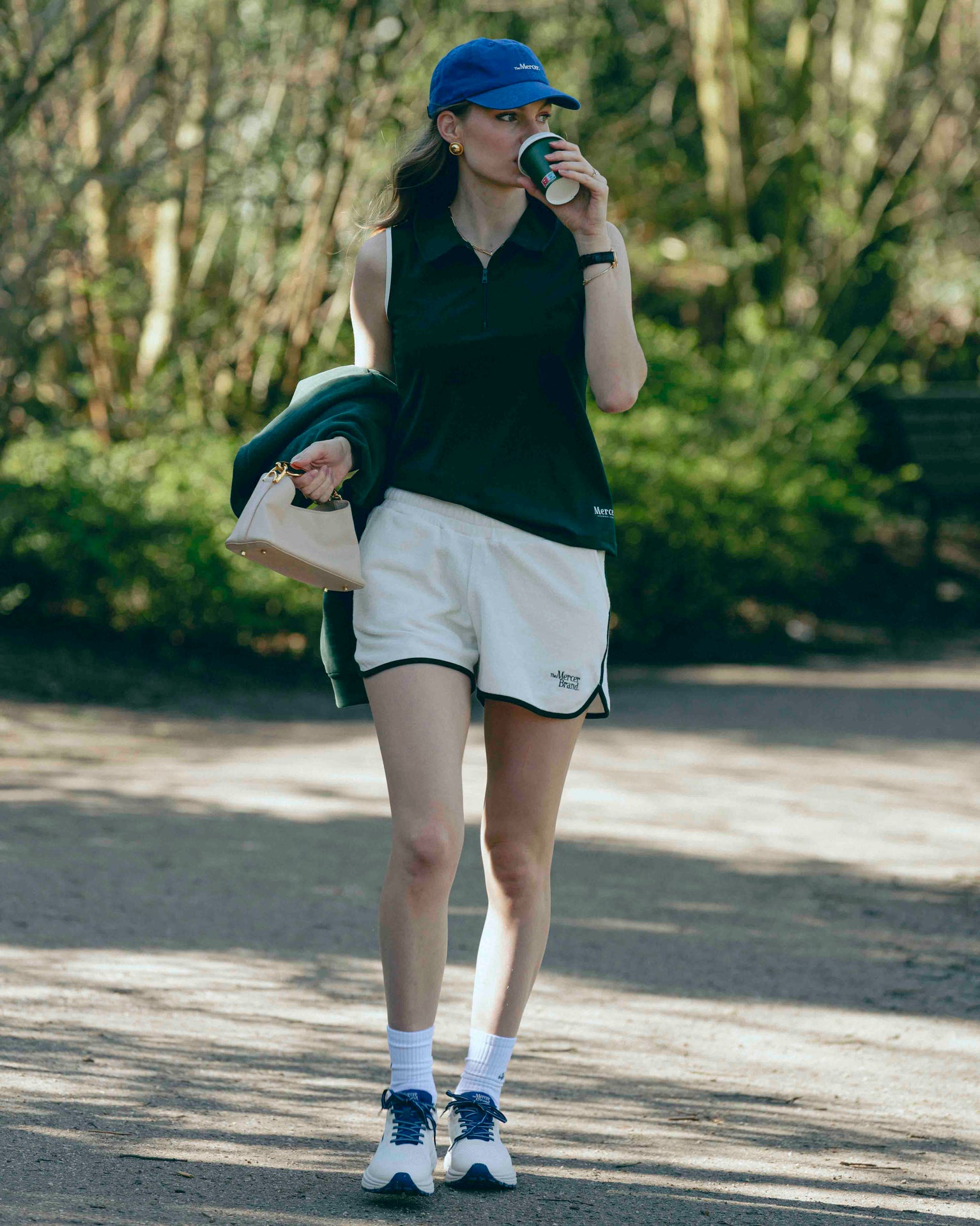 A woman is wearing the Mercer Tennis polo with The shorts and also matching the Mercer Cap with her The Mercer P22 in white/blue. In her hand she is holding her coffee to go.
