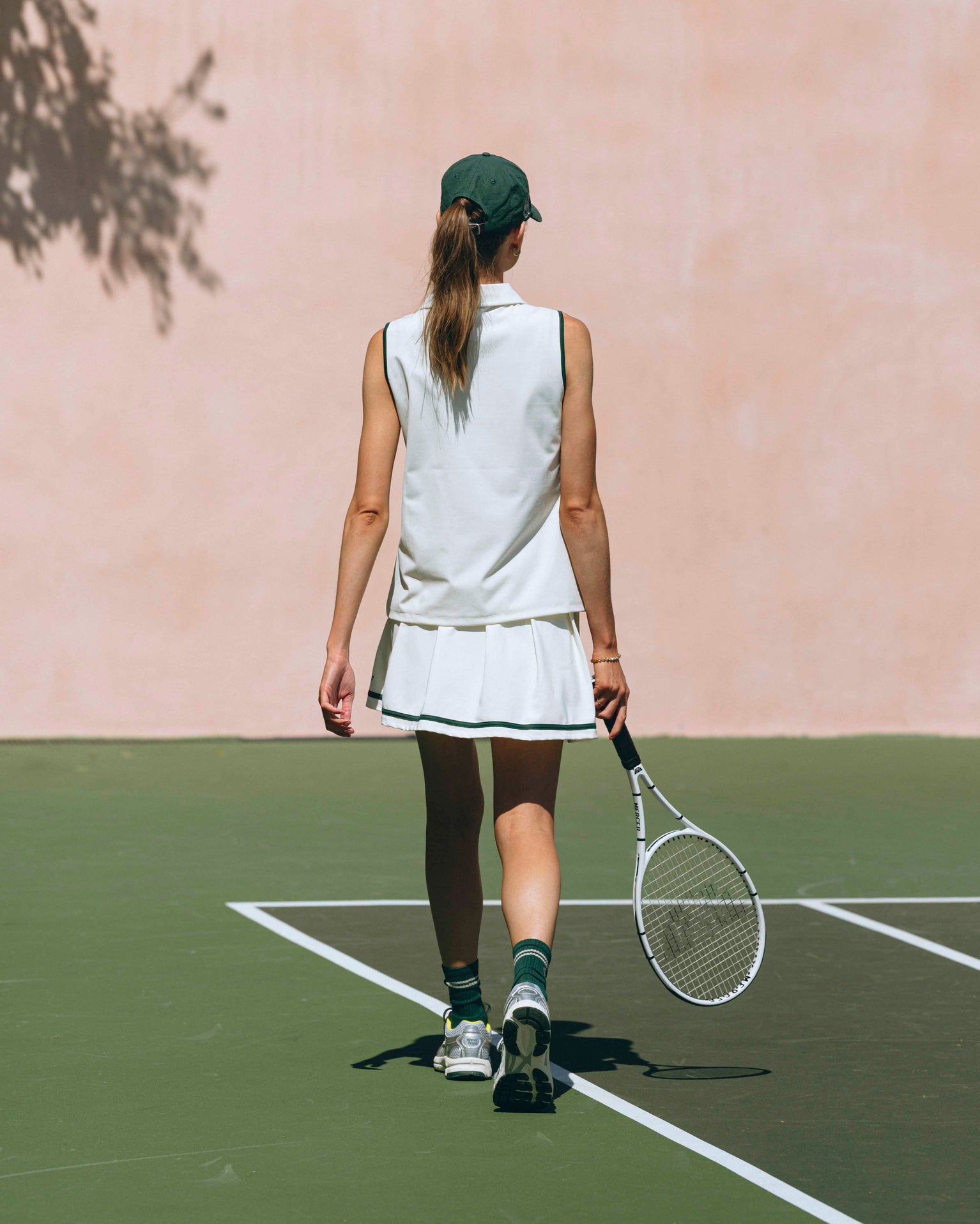 A woman is walking on the court in her The Mercer Tennis Skirt and olo in off white. For shoes she has The Re-Run Evo in silver and a The Mercer Cap in green.