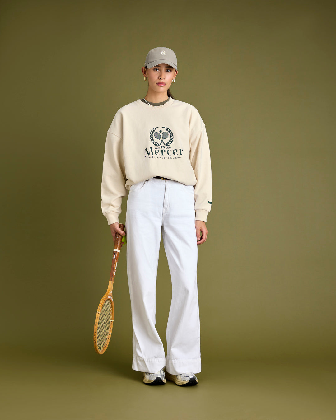 A woman is wearing The Mercer Tennis Sweat in off white with The Mercer 47 Cap and The Re-Run Vibram.